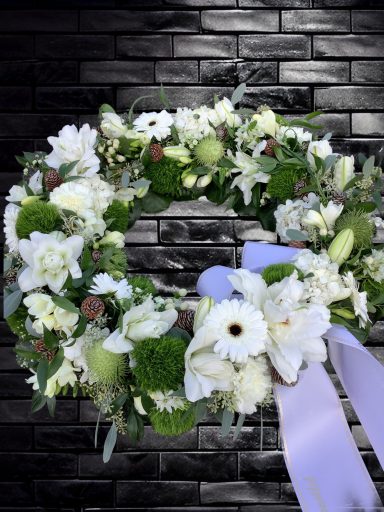 Ein Kranz aus weißen Blumen und grünem Blattwerk, dekoriert mit einem lilanen Band.