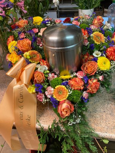 Eine silberne Urne, umgeben von einem Kranz aus bunten Blumen und einem orangefarbenen Band.